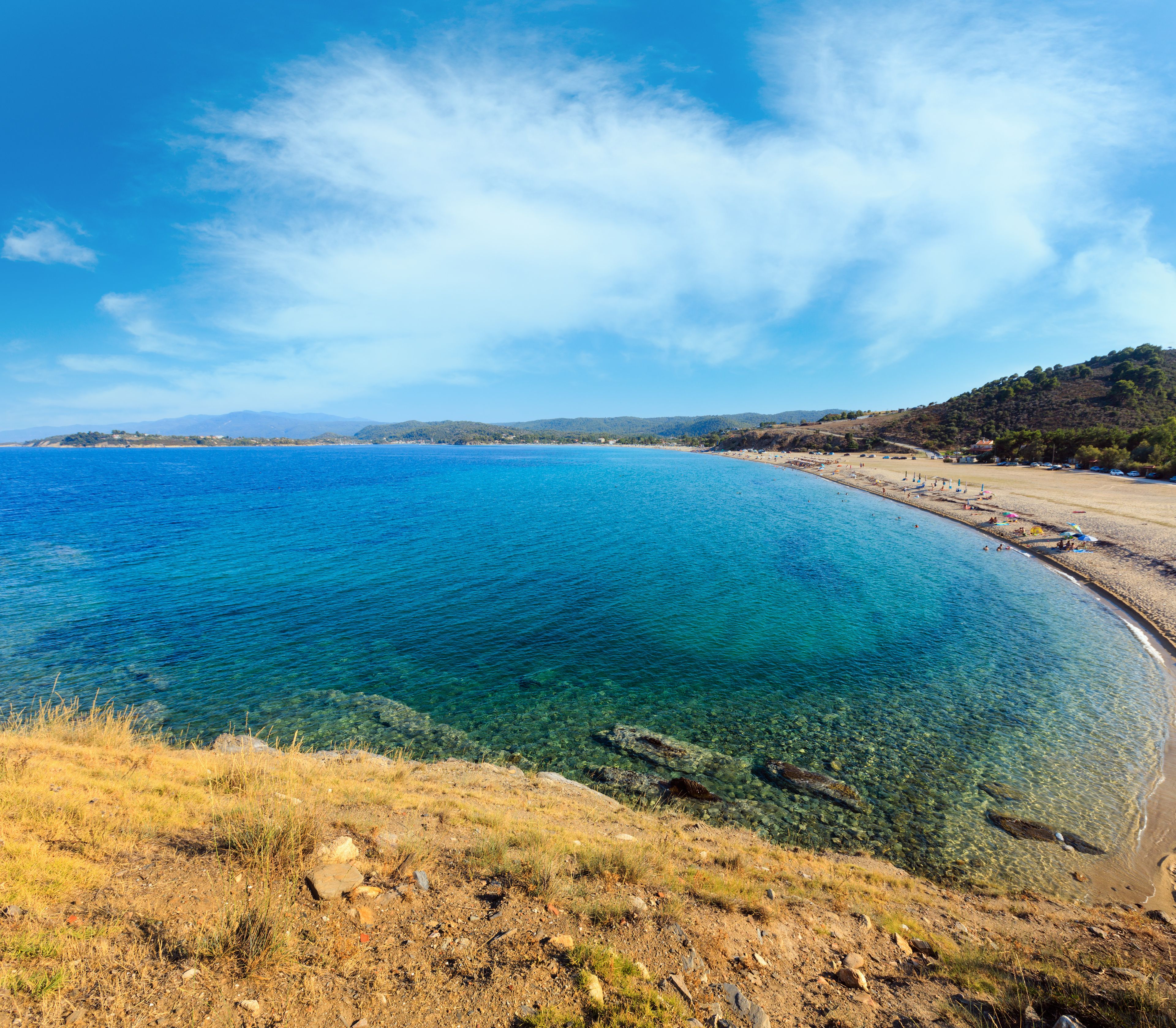 A coastal landscape with clear blue waters, a sandy beach, and a hilly backdrop.