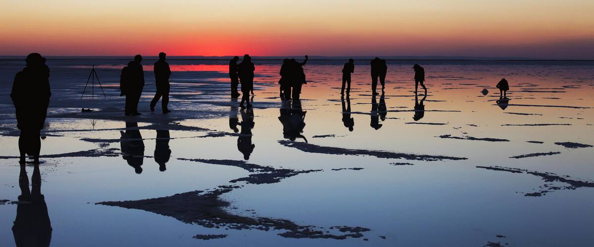 Silhouettes of people standing on a reflective wet surface at sunset.