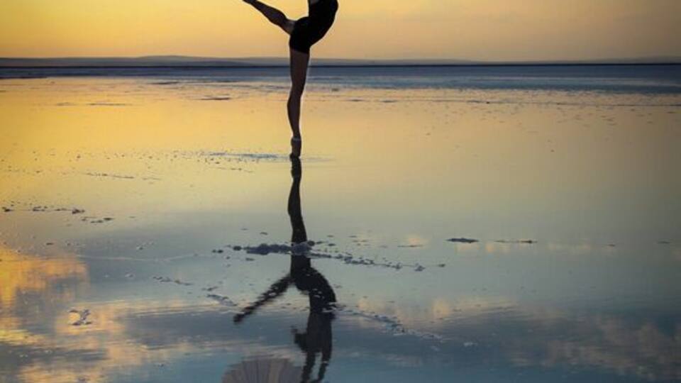 A person balancing on one leg with an umbrella on a reflective beach at sunset.