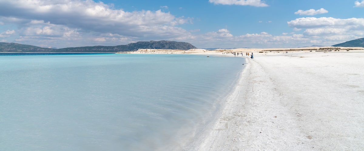 A beach with clear turquoise water, white sand, and distant mountains under a partly cloudy sky.