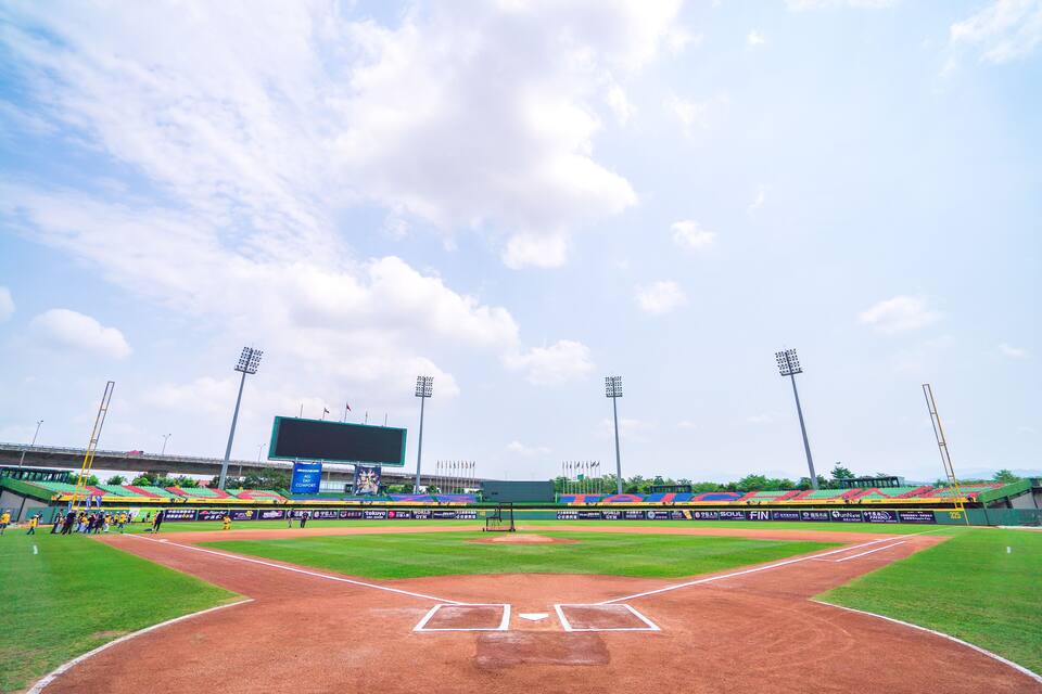 Un estadio de béisbol con infield de tierra, jardín verde y un marcador grande.