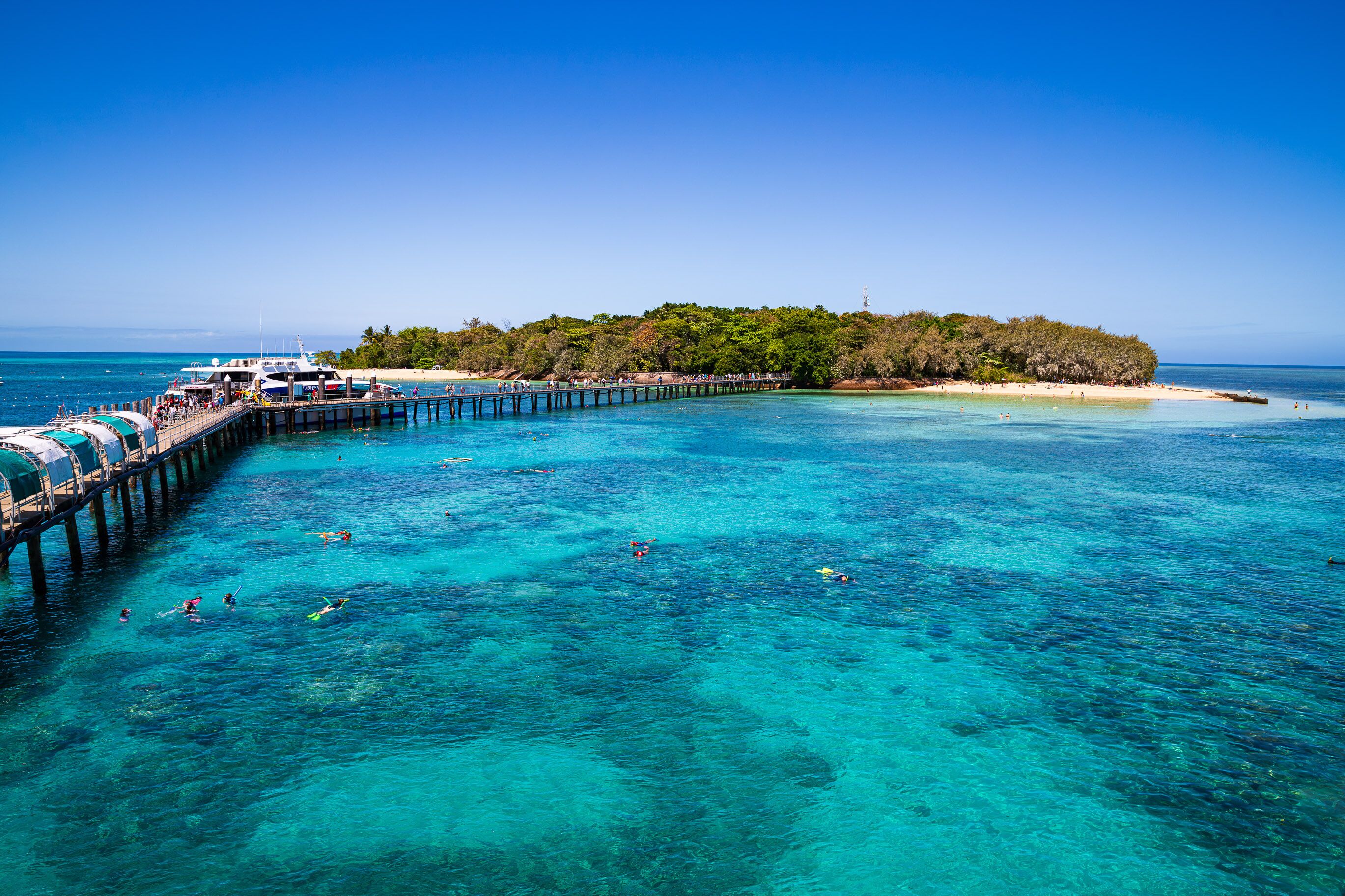 A clear turquoise sea, a pier extending into the water, and a small island with dense vegetation.