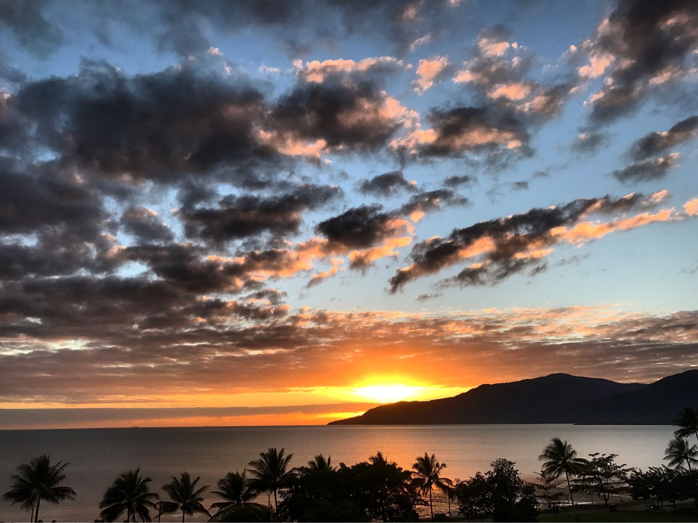 A sunset over a calm sea with palm trees and mountains in the background.