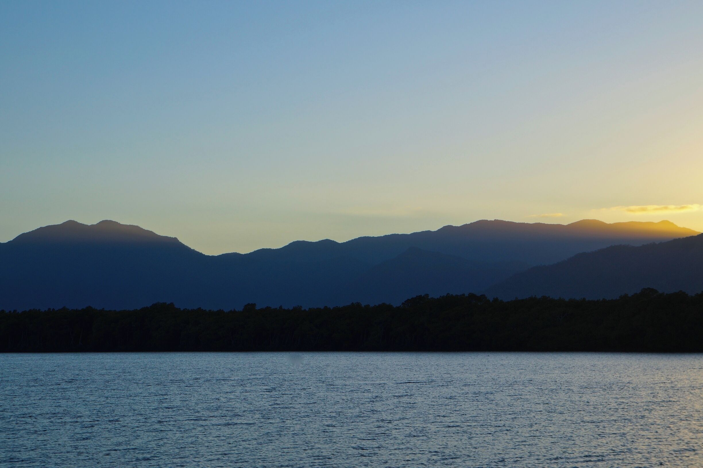A mountain range silhouetted against a sunset, with a calm lake in the foreground.