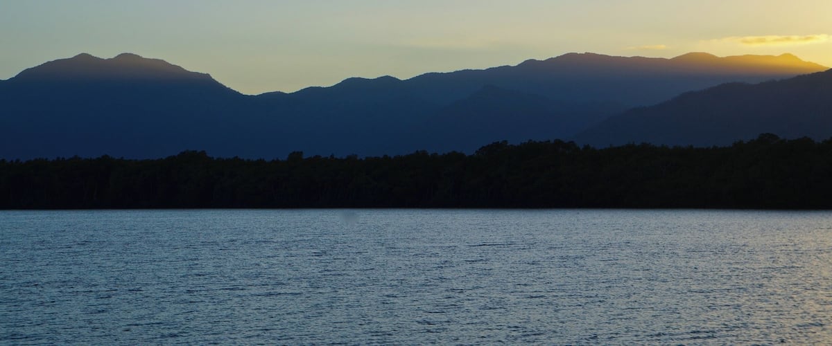 A mountain range silhouetted against a sunset, with a calm lake in the foreground.