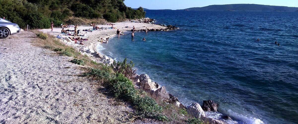 A pebble beach with people swimming and sunbathing, surrounded by greenery and the sea.