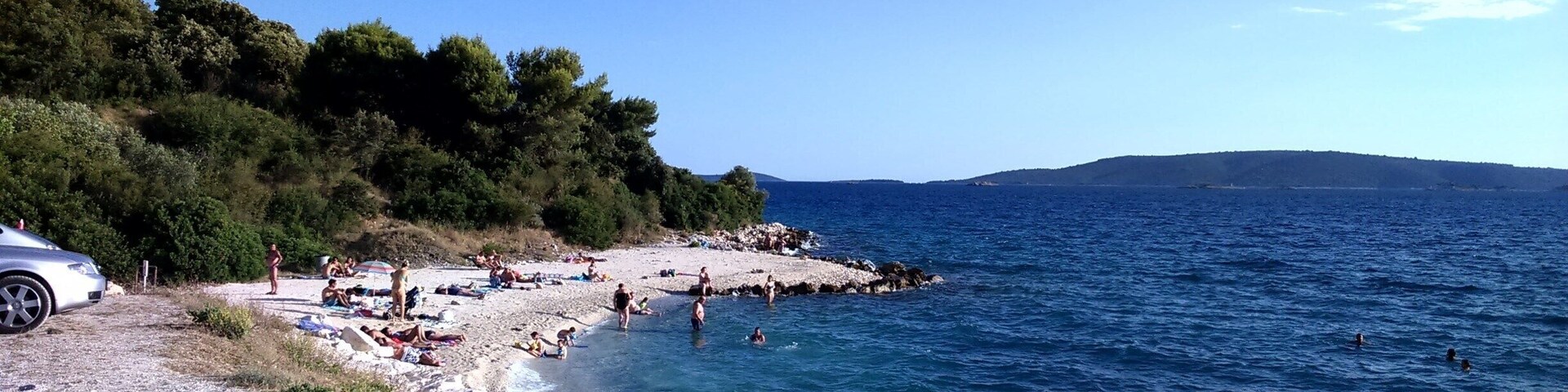A pebble beach with people swimming and sunbathing, surrounded by greenery and the sea.