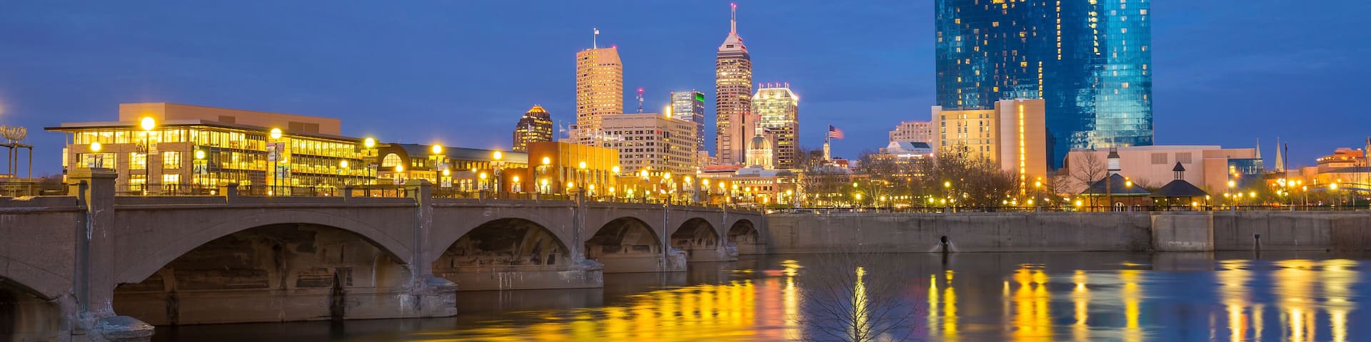 A city skyline at dusk with illuminated buildings, a bridge, and a calm river reflecting the lights.
