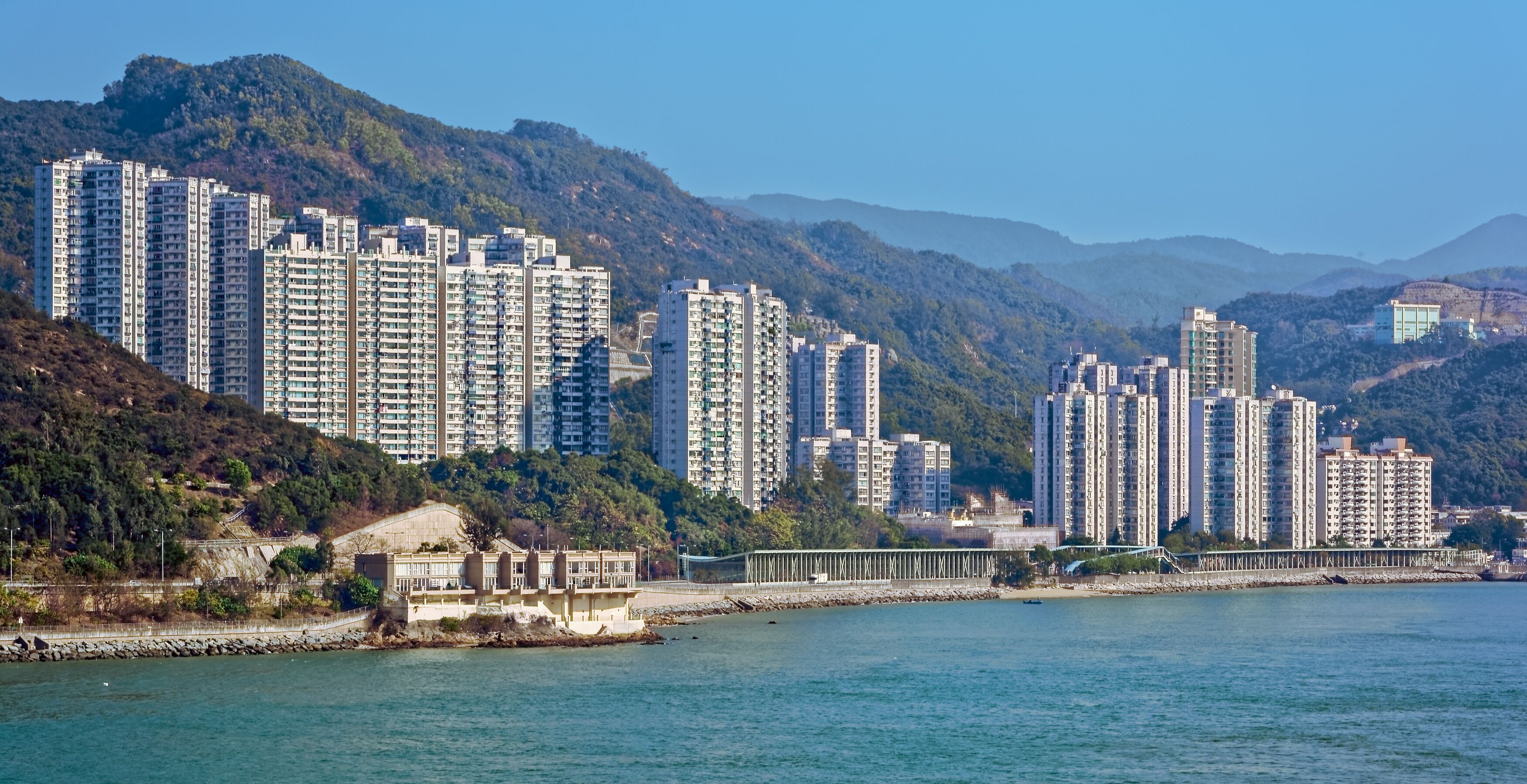 A coastal cityscape with high-rise residential buildings, a bridge, and a mountainous backdrop.