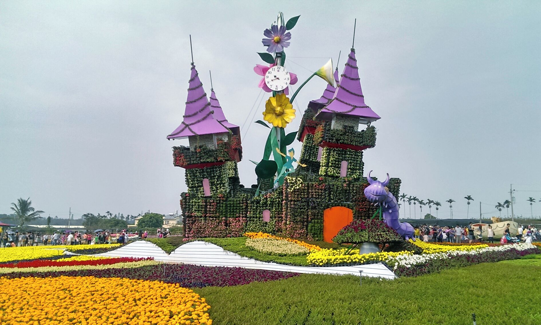A castle-like structure with purple roofs and a clock, surrounded by colorful flower beds.