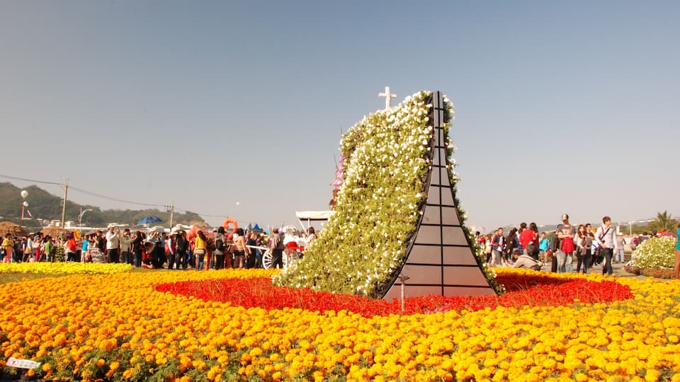 A large crowd gathered around a flower arrangement shaped like a church steeple with a cross on top, surrounded by vibrant yellow and red flowers.