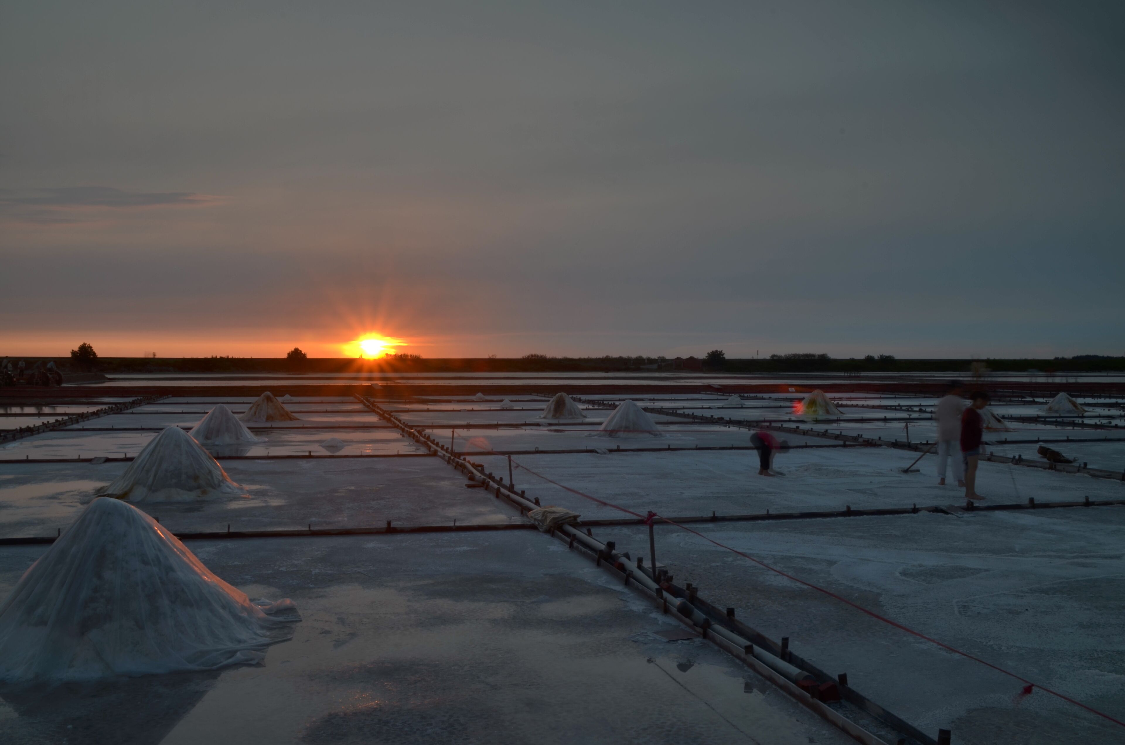 A salt flat at sunset with people working and large white mounds.