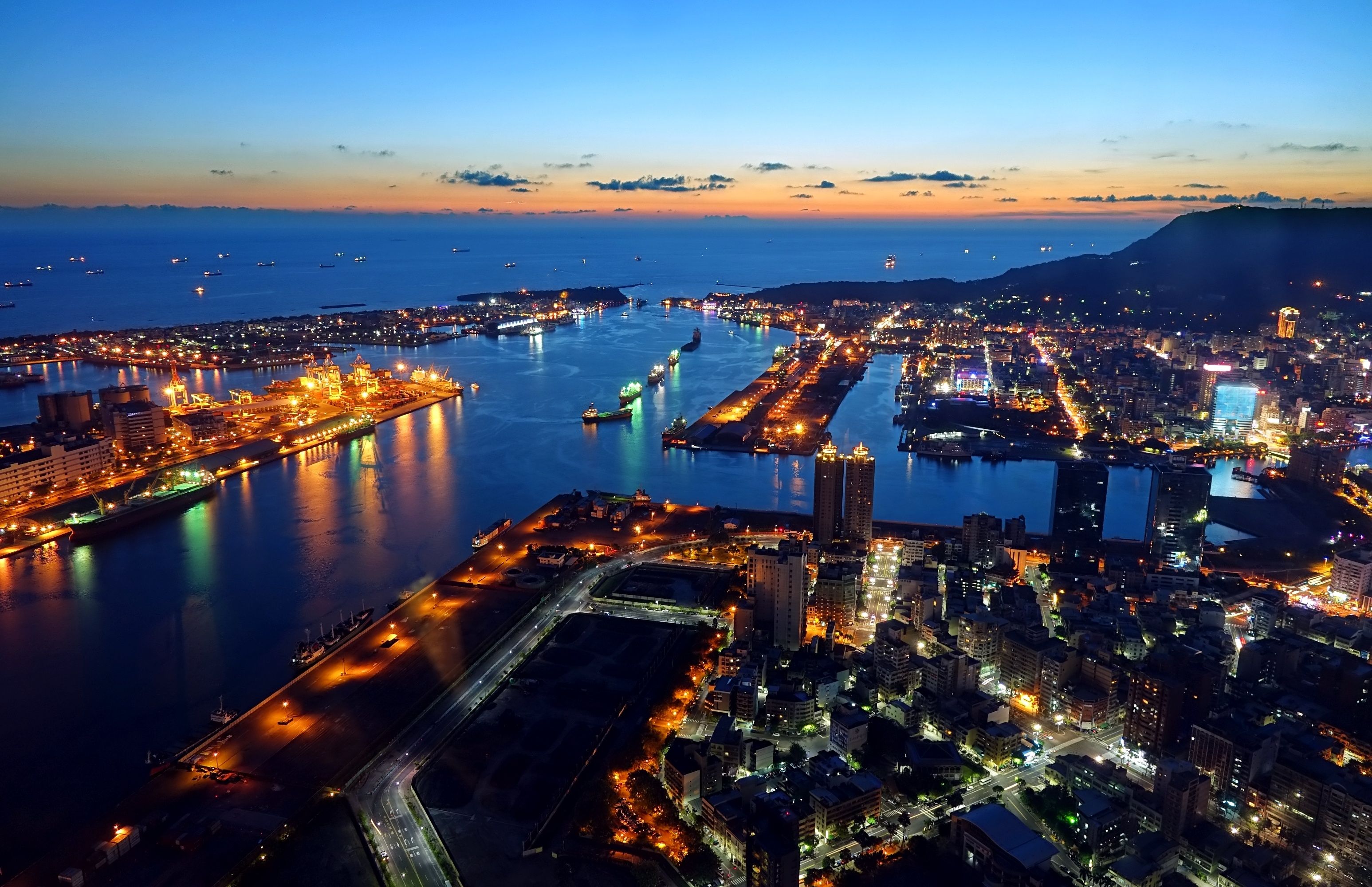A coastal cityscape at dusk with illuminated buildings, a harbor, and a mountain in the background.