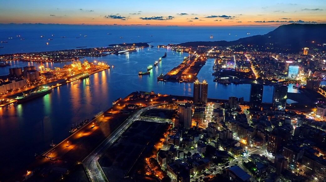 A coastal cityscape at dusk with illuminated buildings, a harbor, and a mountain in the background.