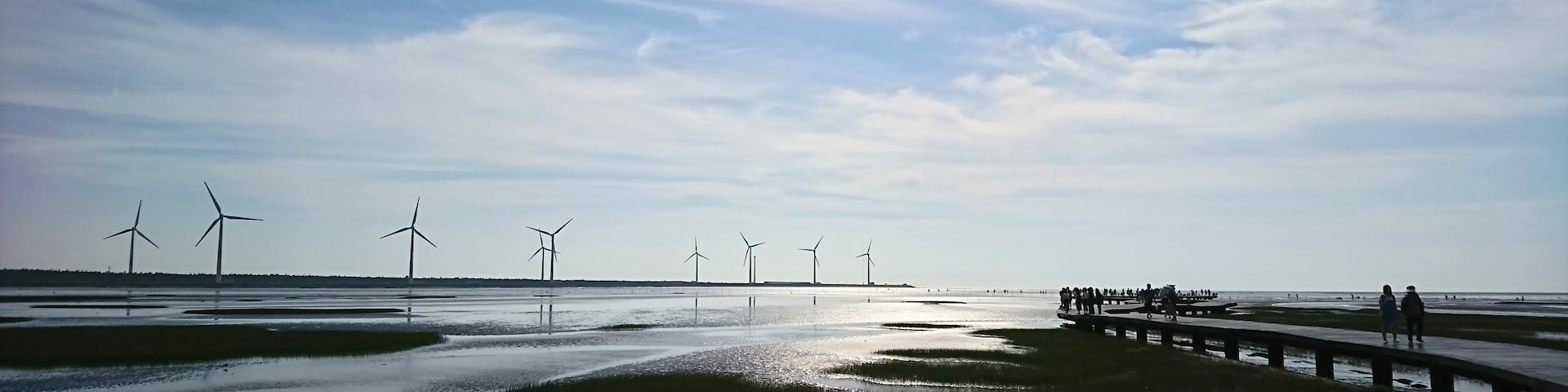 Paisagem costeira com turbinas eólicas e um cais com pessoas.
