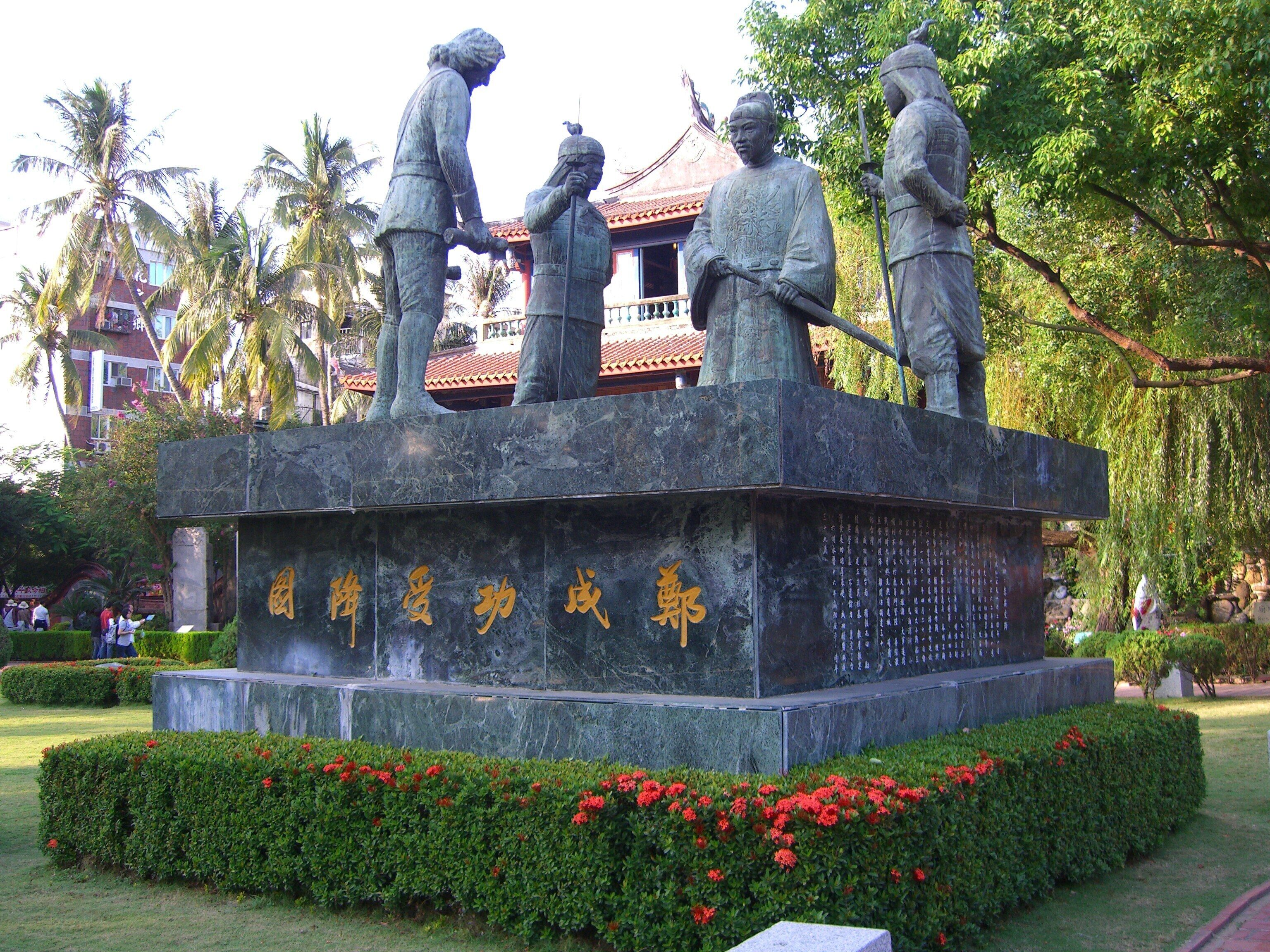 A statue of four figures on a pedestal with inscriptions, surrounded by a garden with red flowers and palm trees.
