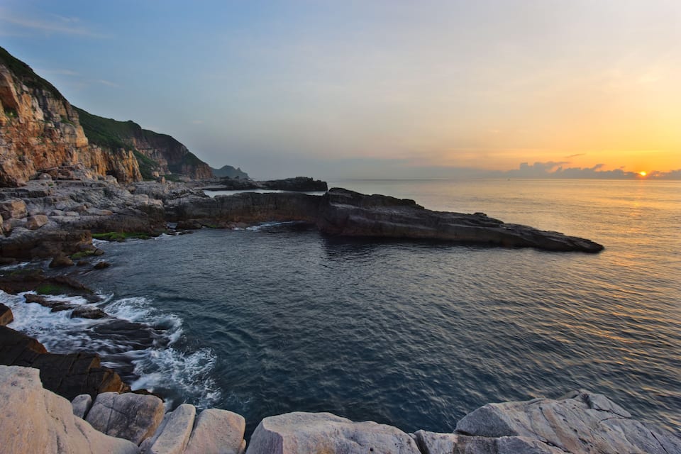 A coastal landscape with rocky cliffs and calm sea at sunset.