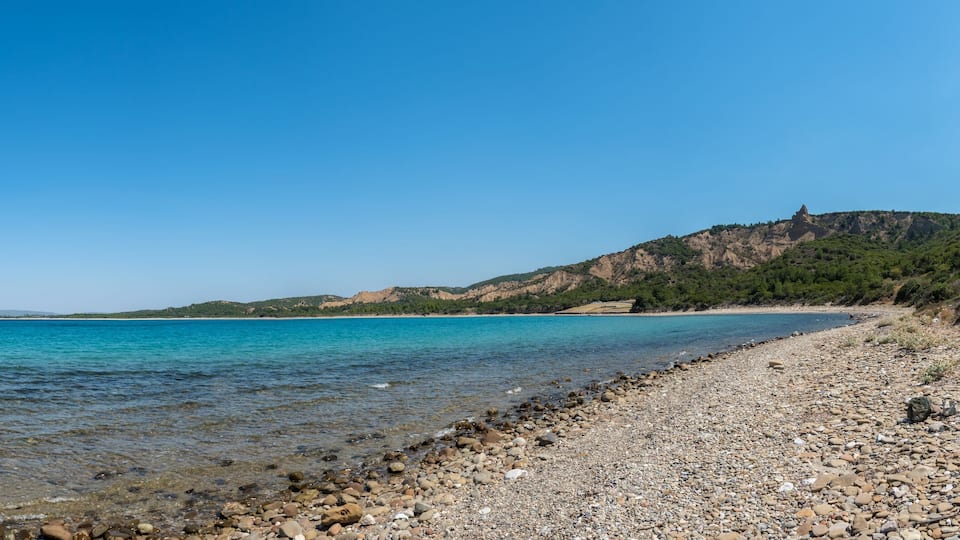 A pebble beach with clear turquoise water, a rocky shoreline, and a hill with sparse vegetation in the background.