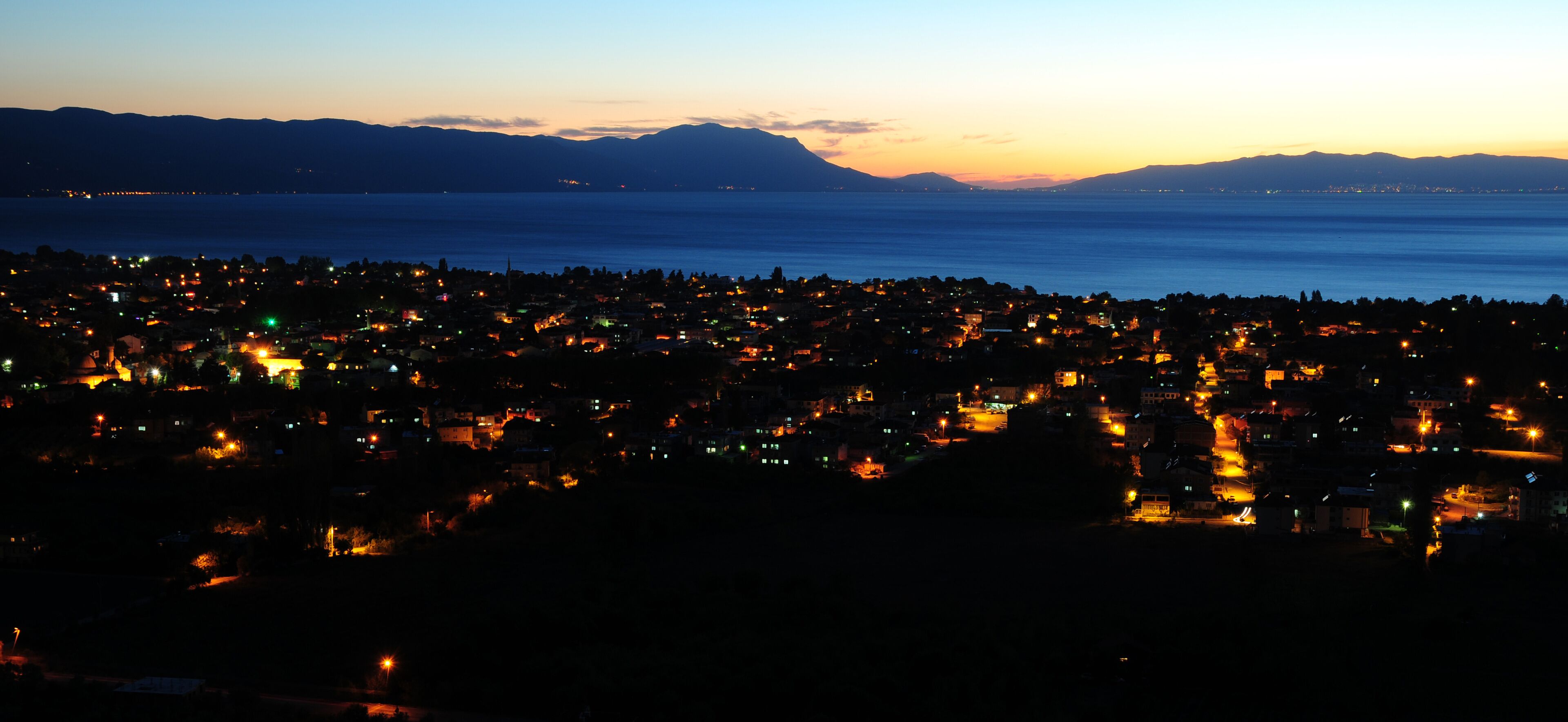 A coastal city at dusk with a view of the sea and mountains in the background.