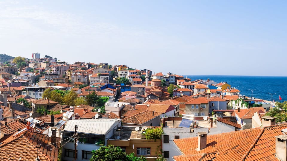 A coastal town with terracotta-roofed buildings, a clear blue sea, and a hilly backdrop.
