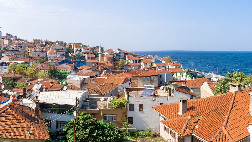A coastal town with terracotta-roofed buildings, a clear blue sea, and a distant mountain range.