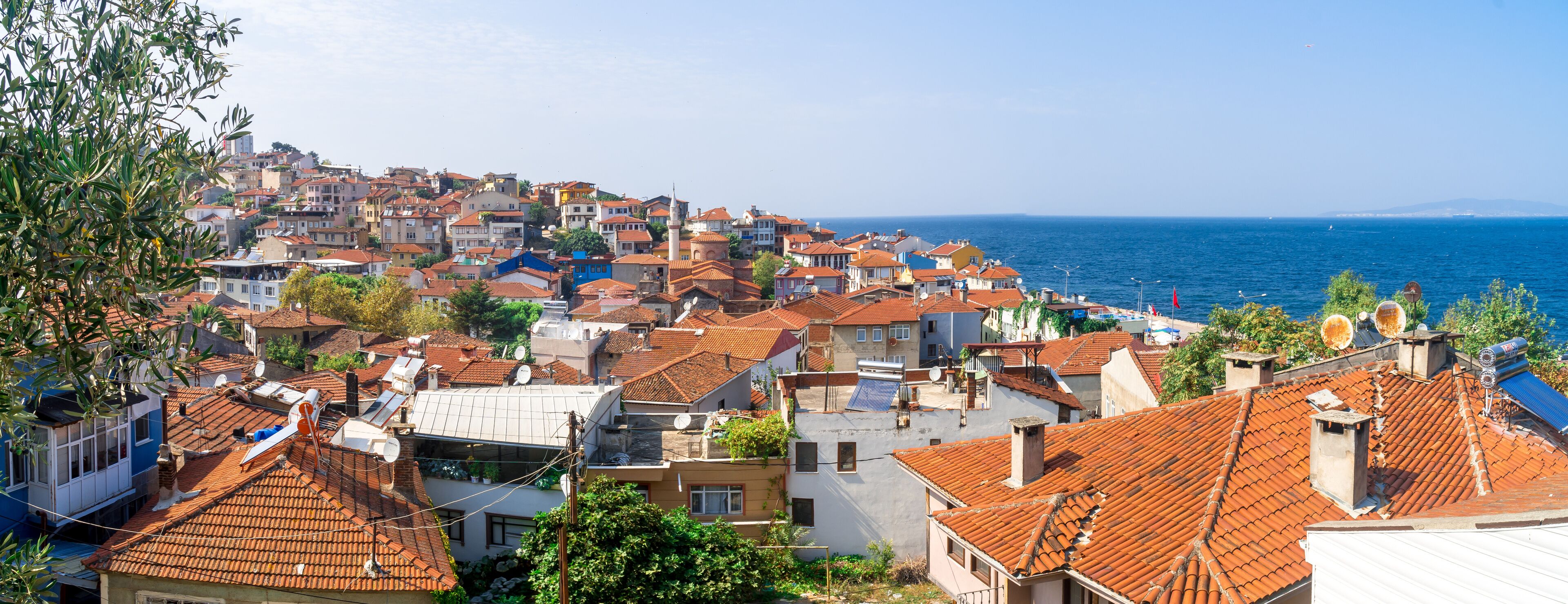 A coastal town with terracotta-roofed buildings, a clear blue sea, and a distant mountain range.