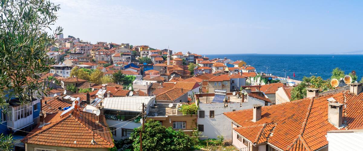 A coastal town with terracotta-roofed buildings, a clear blue sea, and a distant mountain range.