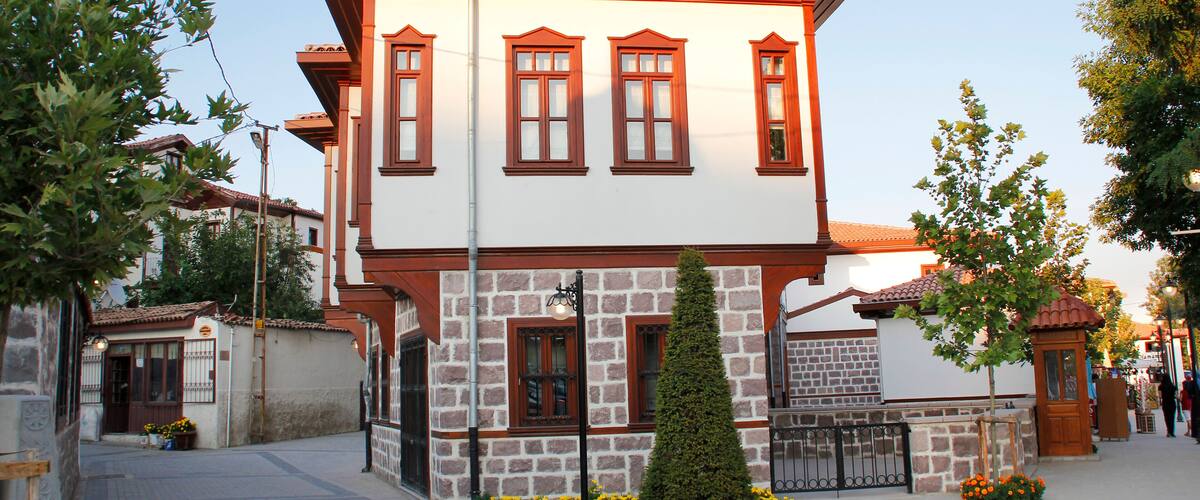 A two-story building with a white facade and red accents, surrounded by a cobblestone street and flowering plants.