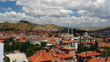 A cityscape with red-roofed buildings, a mosque with two minarets, and a mountainous backdrop under a partly cloudy sky.