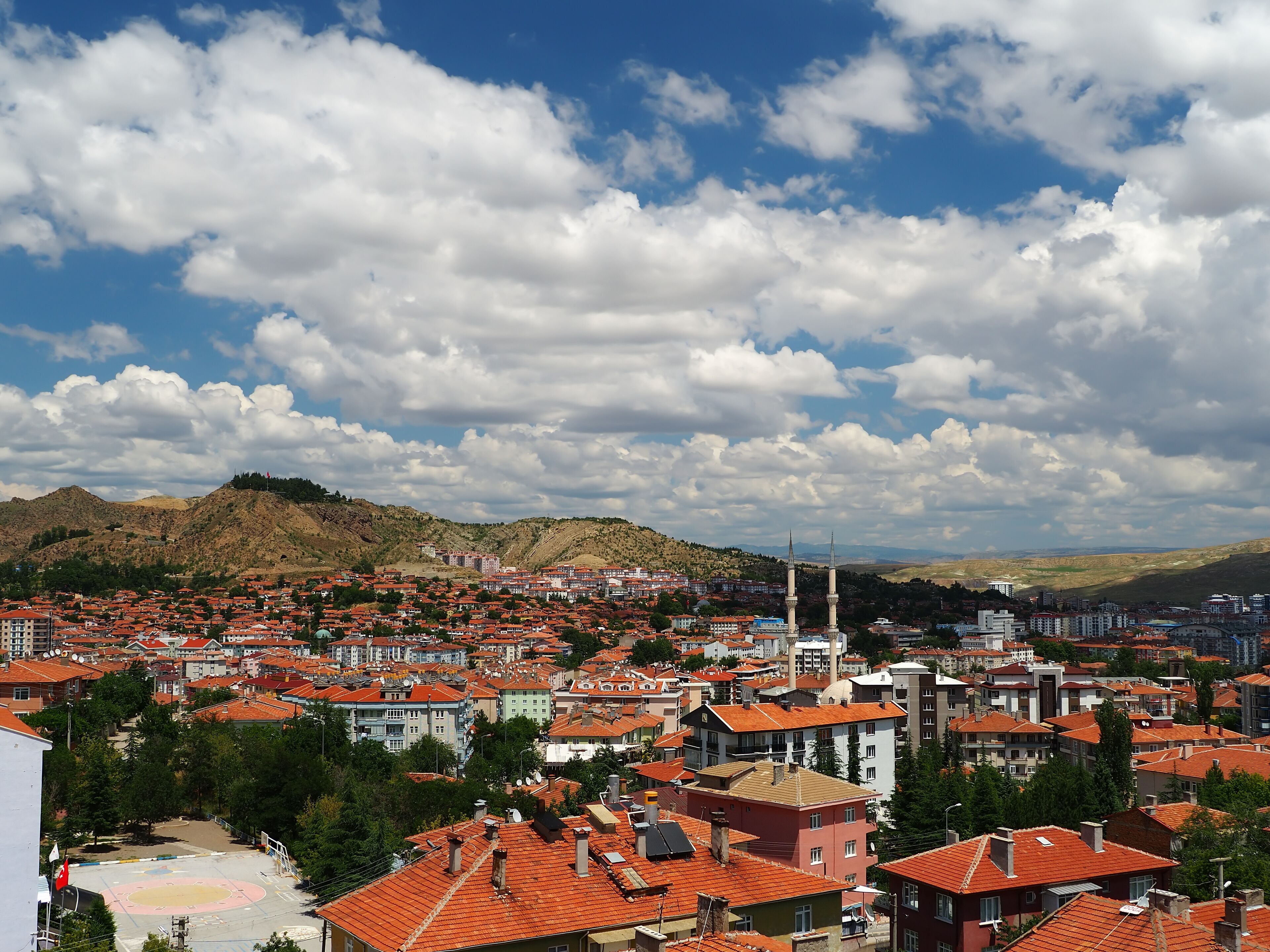 A cityscape with red-roofed buildings, a mosque with two minarets, and a mountainous backdrop under a partly cloudy sky.