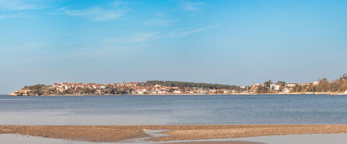 A coastal town with red-roofed buildings, a pebbly beach, and a clear blue sky.