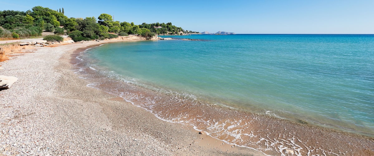 A pebble beach with clear turquoise water, a sandy shore, and a forested hill in the background.
