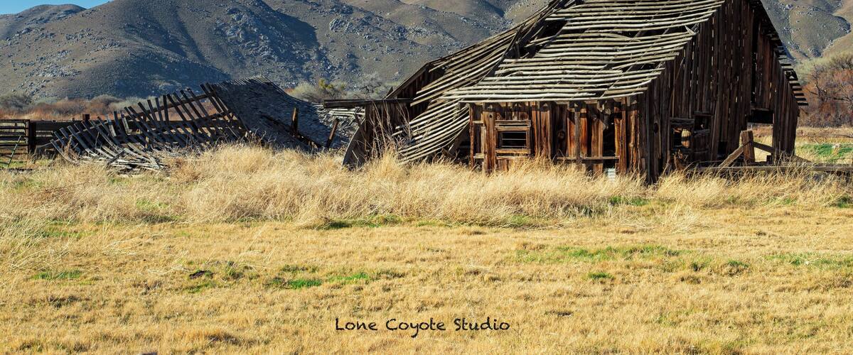 A dilapidated wooden barn with a fence in a grassy field, surrounded by mountains.
