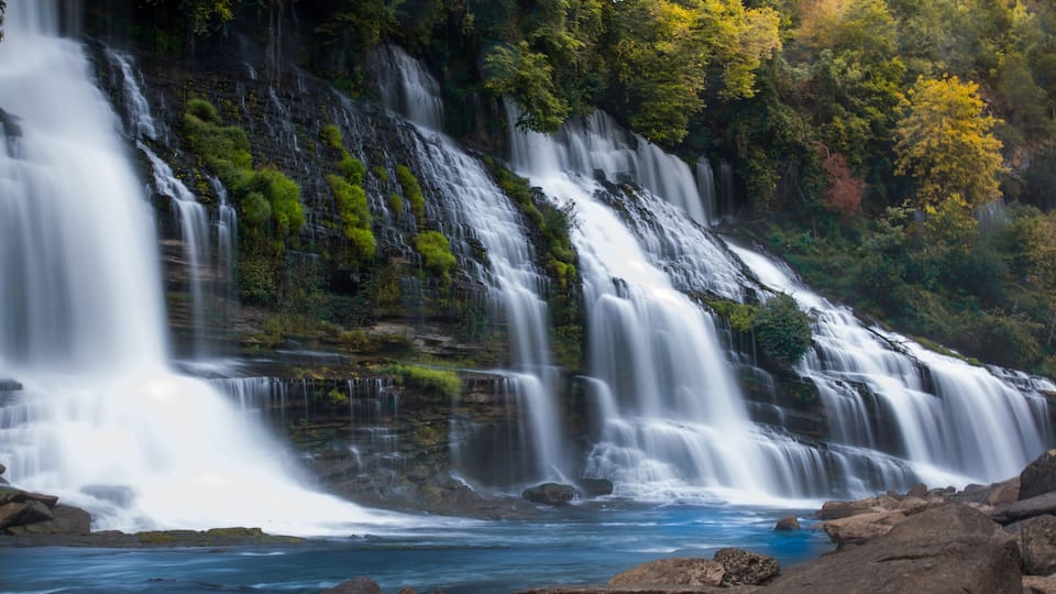 A multi-tiered waterfall with cascading water over rocks, surrounded by lush greenery and trees.