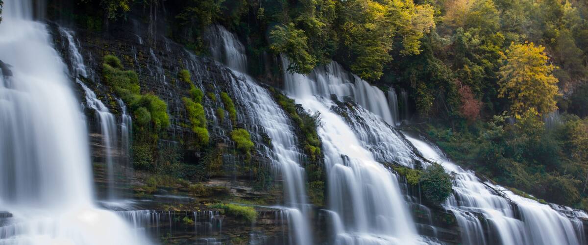 A multi-tiered waterfall with cascading water over rocks, surrounded by lush greenery and trees.