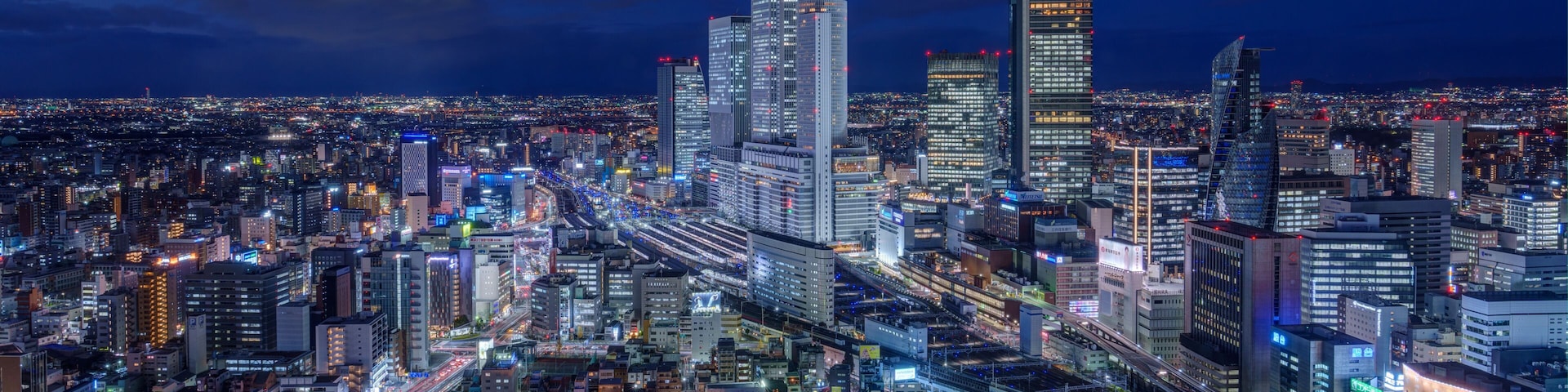 Paisagem urbana noturna com arranha-céus iluminados, traçado urbano denso e uma rede de estradas e autoestradas.