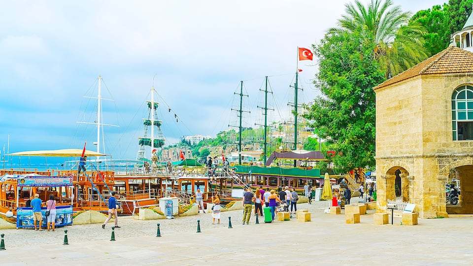 A bustling harbor with sailing ships, a historic stone building, and a clear blue sky.