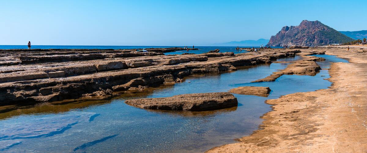 A rocky beach with clear water and a mountain in the background.