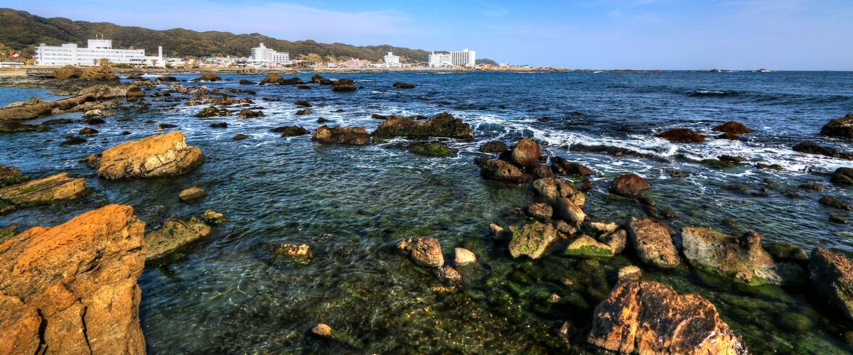A rocky shoreline with clear water, a distant cityscape, and mountains in the background.