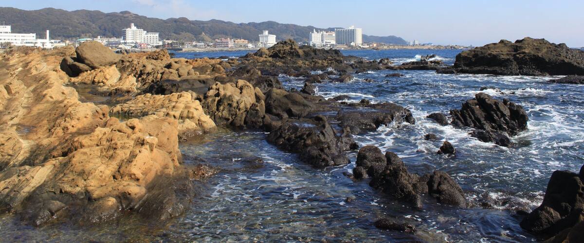 Litoral rochoso com ondas quebrando nas pedras, céu azul límpido e edifícios ao fundo.