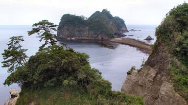 A coastal landscape with a prominent rocky cliff, dense green foliage, and a calm sea.