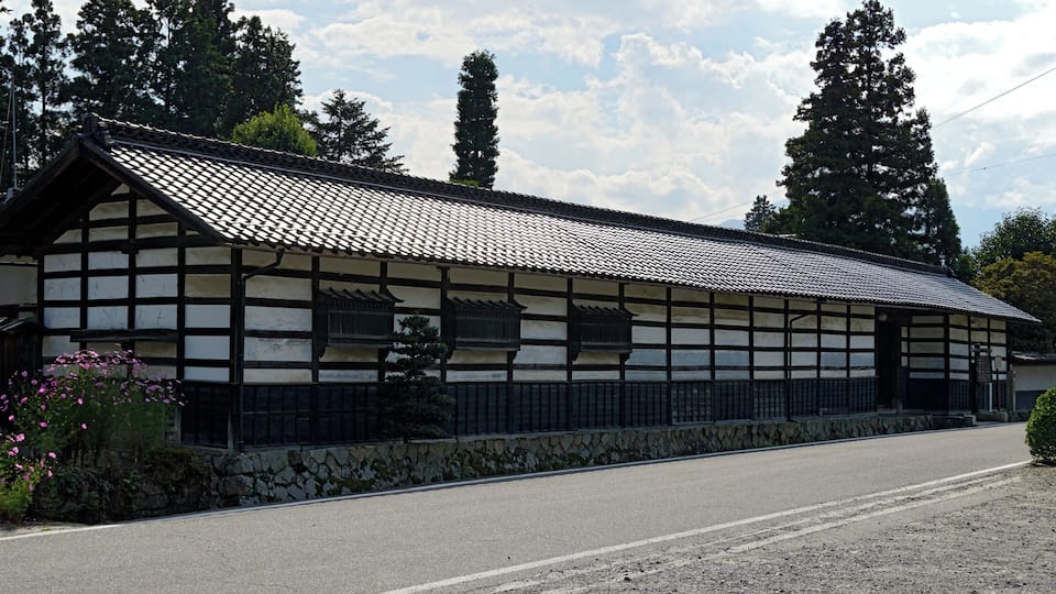 Il s’agit d’un bâtiment japonais traditionnel, doté d’un toit en tuiles et de murs en bois, situé au bord d’une route bordée d’un mur de pierres et d’un petit arbuste.