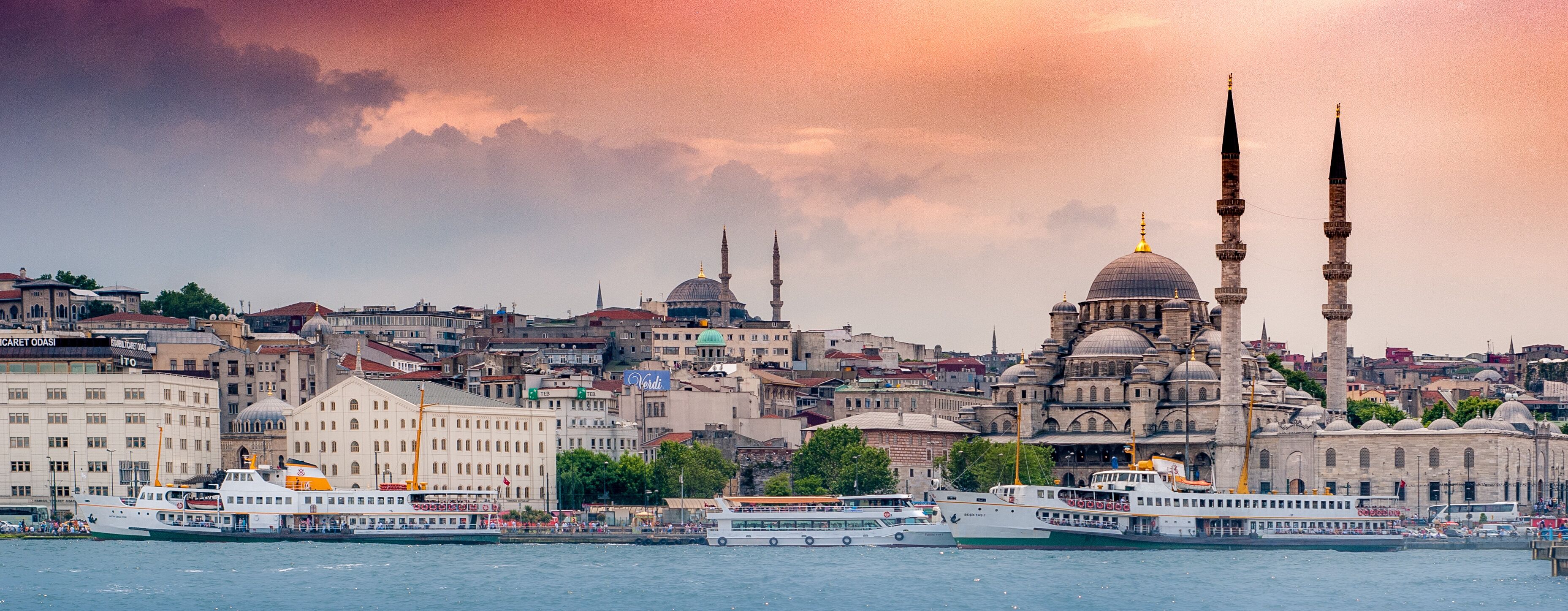 Paisagem urbana com uma mesquita em destaque, com dois minaretes e uma cúpula grande, uma orla marítima com barcos e um cenário urbano denso ao fundo.