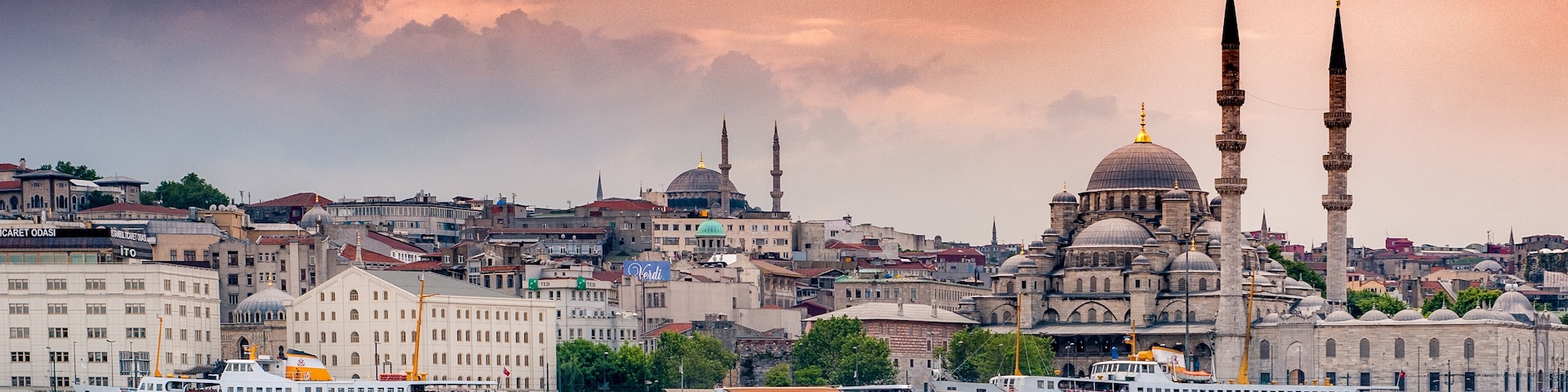 Paisagem urbana com uma mesquita em destaque, com dois minaretes e uma cúpula grande, uma orla marítima com barcos e um cenário urbano denso ao fundo.