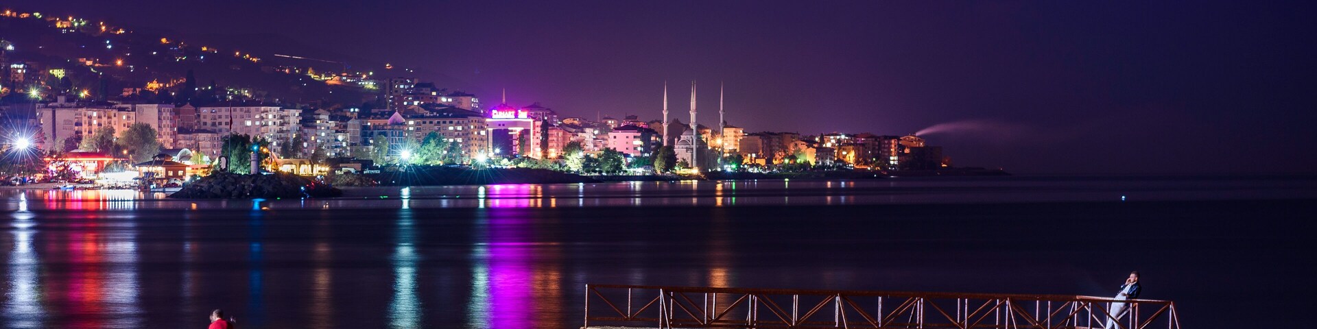 Paisaje costero nocturno con edificios iluminados, un muelle y una mezquita con dos minaretes.