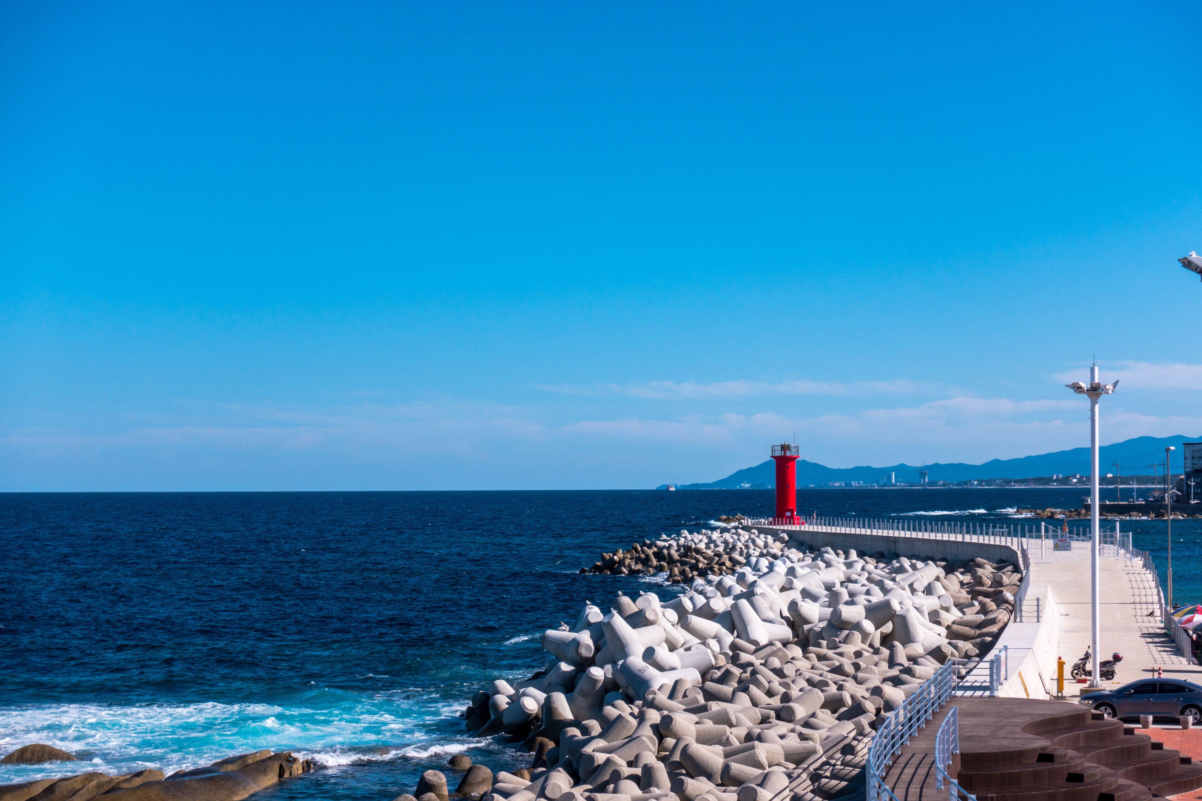 A rocky pier with a red lighthouse, waves crashing against the rocks, and a clear blue sky.
