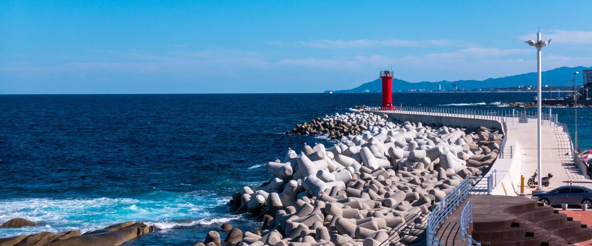 A rocky pier with a red lighthouse, waves crashing against the rocks, and a clear blue sky.