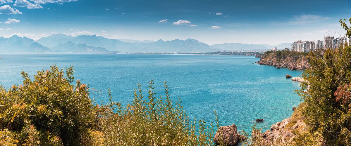 A coastal landscape with a clear blue sea, mountains in the background, and a city skyline.