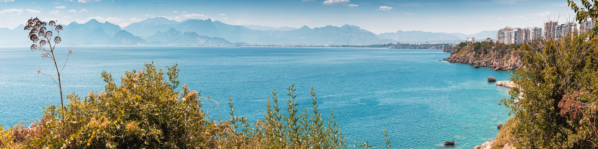 A coastal landscape with a clear blue sea, mountains in the background, and a city skyline.