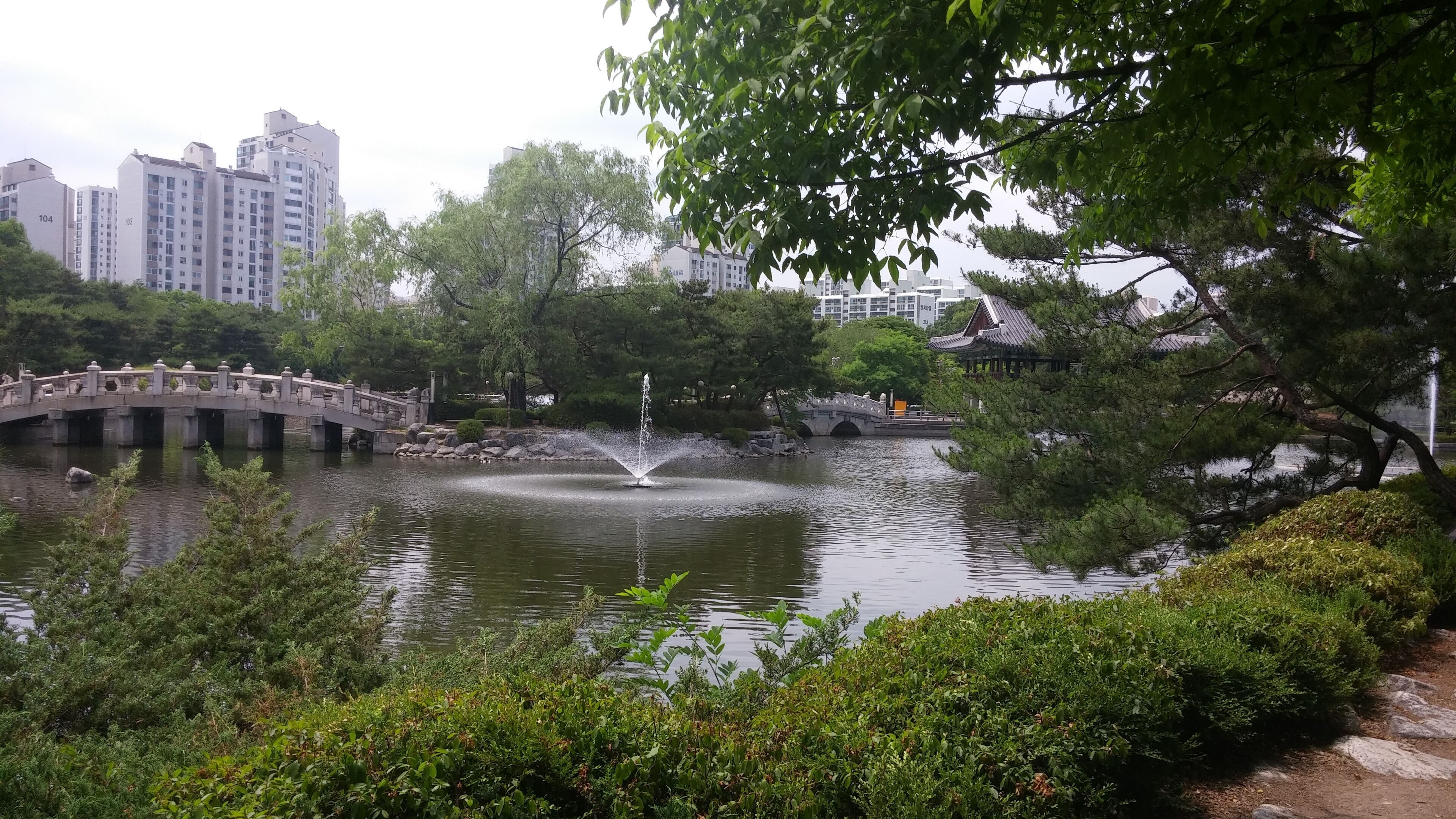 A serene park with a pond, a stone bridge, and traditional architecture in the background.